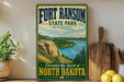 A vintage-style sign for Fort Ransom State Park, featuring a scenic landscape with a river and mountains, is displayed on a wooden shelf next to a wooden cutting board and some lemons.