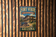 A vintage-style metal sign for Fort Verde State Historic Park in Arizona, featuring a building and a wagon wheel, mounted on a wooden fence.