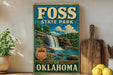 A vintage-style sign for Foss State Park in Oklahoma, featuring a waterfall and lush greenery, is displayed on a shelf next to a wooden cutting board and some lemons.