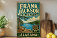 A vintage-style sign for Frank Jackson State Park in Alabama, featuring a scenic landscape with a lake and pine trees.