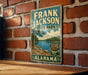 A vintage-style sign for Frank Jackson State Park in Alabama, featuring a scenic landscape with a lake and mountains.