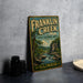 A vintage Franklin Creek sign from Illinois, featuring a scenic landscape with a river and mountains, is displayed on a gray countertop next to some black camera lenses.