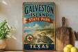 A vintage-style sign for Galveston Island State Park in Texas, featuring a turtle and beach scene, is displayed on a shelf next to a wooden cutting board and some lemons.