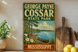 A vintage-style sign for George Payne Cossar State Park in Mississippi, featuring a scenic image of a lake, trees, and a canoe.