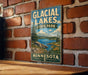A vintage-style metal sign for Glacial Lakes State Park in Minnesota, featuring a scenic landscape with a lake, mountains, and trees.