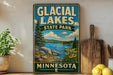 A vintage-style sign for Glacier Lakes State Park in Minnesota, featuring a scenic landscape with a lake, mountains, and pine trees.