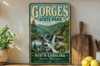 A vintage-style metal sign for Gorges State Park in North Carolina, featuring a scenic mountain landscape with a waterfall. The sign is displayed on a wooden shelf, surrounded by various kitchen utensils and a potted plant.