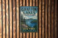 A vintage-style metal sign for Grafton Lakes State Park, featuring a scenic landscape with a lake and mountains, mounted on a wooden wall.