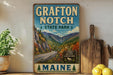 A vintage-style sign for Grafton Notch State Park in Maine, featuring a scenic mountain landscape with a winding road and pine trees.
