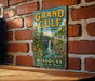A vintage metal sign for Grand Gulf State Park, featuring a scenic illustration of a waterfall and lush greenery, mounted on a brick wall.