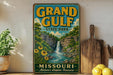 A vintage-style sign for Grand Gulf State Park in Missouri, featuring a waterfall and lush greenery, is displayed on a shelf next to a wooden cutting board and some potted plants.