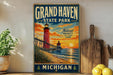 A vintage-style metal sign for Grand Haven State Park in Michigan, featuring a lighthouse and a sunset over the ocean.
