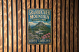 A vintage metal sign for Grandfather Mountain State Park in North Carolina, featuring a scenic mountain landscape with a winding road and pink flowers.