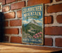 A vintage-style metal sign for Grandfather Mountain State Park in North Carolina, featuring a scenic mountain landscape with a winding road and pink flowers.