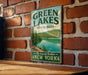 A vintage-style metal sign for Green Lakes State Park in New York, featuring a scenic image of a lake surrounded by pine trees and mountains.