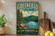 A vintage-style sign for Greenfield State Park in New Hampshire, featuring a scenic landscape with pine trees, a lake, and mountains.