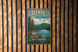 A vintage-style metal sign for Greenfield State Park in New Hampshire, featuring a scenic landscape with pine trees, a lake, and mountains.