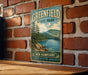 A vintage-style metal sign for Greenfield State Park in New Hampshire, featuring a scenic landscape with pine trees, a lake, and mountains.
