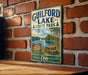 A vintage metal sign for Guilford Lake State Park, featuring a scenic image of a lake, picnic table, and trees, mounted on a brick wall.