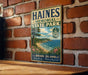 A vintage metal sign for Haines Memorial State Park in Rhode Island, featuring a scenic coastal landscape with a beach, ocean, and mountains.