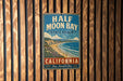 A vintage metal sign for Half Moon Bay State Beach, California, featuring a coastal landscape with a beach, ocean, and mountains.