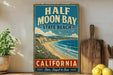 A vintage-style metal sign for Half Moon Bay State Beach in California, featuring a beach scene with mountains and a blue sky.