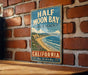A vintage-style metal sign for Half Moon Bay, California, mounted on a brick wall.