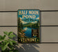 A vintage-style metal sign for Half Moon Pond State Park in Vermont, featuring a scenic landscape with a lake, mountains, and pine trees.