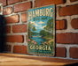 A vintage metal sign for Hamburg State Park in Georgia, featuring a scenic landscape with a river, trees, and a waterfall.