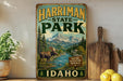 A vintage-style metal sign for Harriman State Park in Idaho, featuring a moose and a mountain landscape.