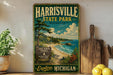 A vintage-style sign for Harrisville State Park in Michigan, featuring a scenic landscape with a lake, pine trees, and a beach.