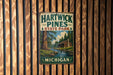 A vintage metal sign for Hartwick Pines State Park in Michigan, featuring a scenic illustration of a cabin surrounded by pine trees and a stream.