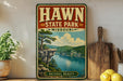 A vintage-style sign for Hawn State Park in Missouri, featuring a scenic landscape with a lake and mountains.