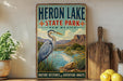 A vintage-style poster forHERON LAKE STATE PARK, NEW MEXICO, featuring a large heron in a grassy landscape with mountains in the background.