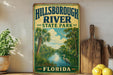 A vintage-style sign for Hillsborough River State Park in Florida, featuring a serene landscape with palm trees, a heron, and a river.
