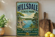 A vintage-style metal sign for Hillsdale State Park in Kansas, featuring a scenic landscape with a lake and mountains.
