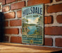 A vintage metal sign for Hillsdale State Park in Kansas, featuring a scenic landscape with a lake and mountains.
