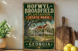 A vintage-style sign for Hofwyll Broadfield Plantation State Park in Georgia, featuring a house and palm trees, is displayed on a shelf next to a wooden cutting board and some lemons.