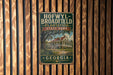A vintage-style metal sign for Hofwyll Broadfield Plantation State Park in Georgia, featuring a large illustration of a plantation house surrounded by palm trees.