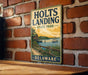 A vintage-style metal sign for Holts Landing State Park in Delaware, featuring a scenic illustration of a boat on a river with a wooden dock and a house in the background.