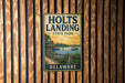 A vintage metal sign for Holts Landing State Park in Delaware, featuring a scenic image of a lake and a dock with a house and a boat.