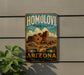 A vintage-style metal sign for Homolovi State Park in Arizona, featuring an illustration of a desert landscape with a large stone structure.
