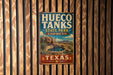 A vintage-style metal sign for Hueco Tanks State Park & Historic Site in Texas, featuring a desert landscape with a cactus and a small pool.
