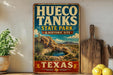 A vintage-style sign for Hueco Tanks State Park & Historic Site in Texas, featuring a desert landscape with a small lake and mountains in the background.