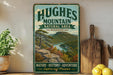 A vintage-style sign for the Hughes Mountain Natural Area in Missouri, featuring a scenic landscape with mountains, trees, and a river.