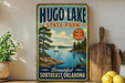 A vintage-style sign for Hugo Lake State Park, featuring a scenic illustration of a lake, a sailboat, and a bird, along with the text "Beautiful Southeast Oklahoma" and "FISH Boat Explore".