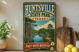 A vintage-style metal sign for the state park called "Huntsville State Park" in Texas, featuring a scenic landscape with a lake, bridge, and pine trees.