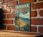 A vintage-style sign for Indiana Dunes State Park, featuring a scenic illustration of the park's landscape with a beach, lake, and mountains.