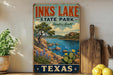 A vintage-style sign for Inks Lake State Park in Texas, featuring a scenic landscape with a lake, mountains, and desert.