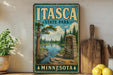 A vintage-style sign for Itasca State Park in Minnesota, featuring a scenic landscape with a river, pine trees, and a monument.